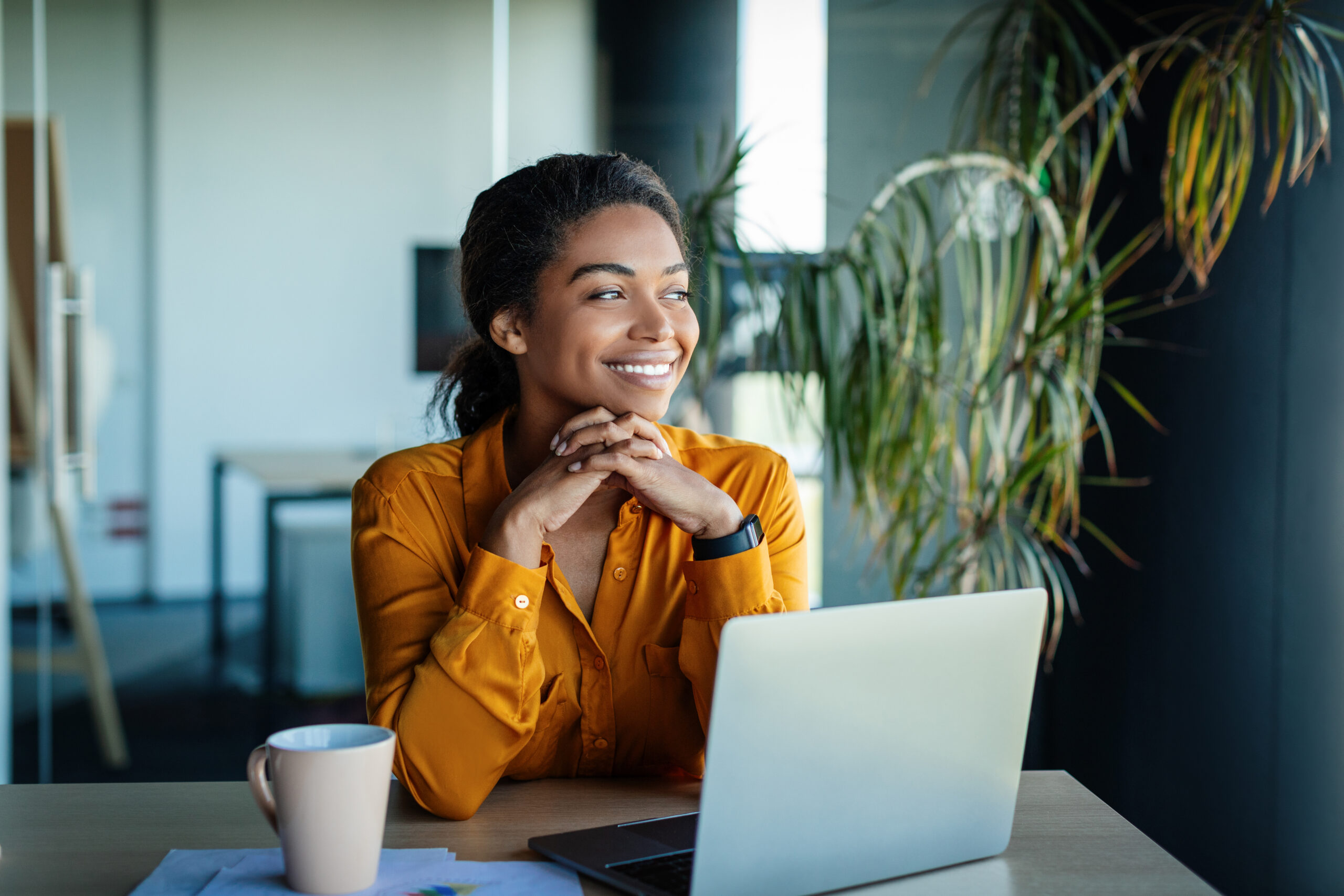 Portrait of dreamy african american businesswoman sitting at desk in office and thinking while working on laptop, looking away with pleased face expression, copy space